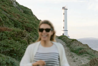 Mujer sonriendo desenfocada en primer plano, con un faro blanco al fondo y vegetación gallega a los lados del camino costero.