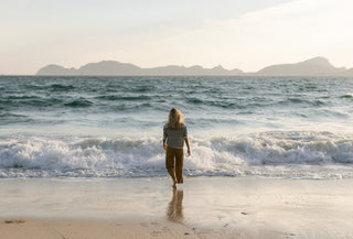Autorregalo: no es capricho, es autocuidado- Mujer frente a la orilla de la playa vestida con jersey y pantalón largo. Al fondo montañas de poca altura.