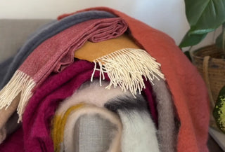 Pila de mantas de mohair y lana en tonos intensos sobre el respaldo de un sofá. Colores de montaña y calor de hogar.