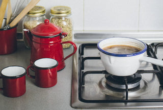 Escena de cocina con cafetera de peltre roja, cazuela de peltre blanca y dos tazas, todo dispuesto para hacer café de pucherete.