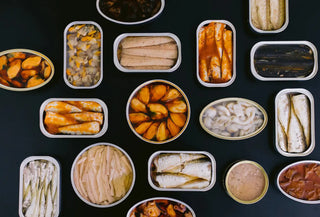 Vista cenital sobre fondo negro de latas de conserva abiertas de mejillones, sardinillas, berberechos y navajas.