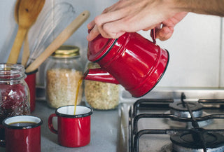 Cafetera y tazas de peltre rojo sirviendo café en una cocina sin plásticos, con tarros de cristal y utensilios de madera reutilizables. Vivir sin plástico: el futuro tiene mucho de pasado Blog Real Fábrica