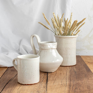 Vista de 3 piezas de cerámica artesana con acabado en esmalte blanco craquelado: vaso con asa, jarra con asa y portacubiertos. El portacubiertos contiene un ramillete de espigas doradas.