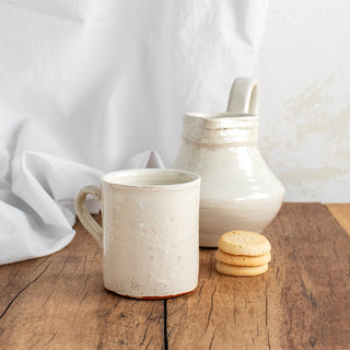 Vista de 2 piezas de cerámica artesana con acabado en esmalte blanco craquelado: vaso con asa y jarra con asa. 