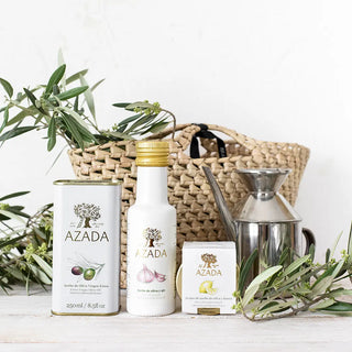 Foto del aceite de ajo, acompañado de otros aceites a su lado, una aceitera clásica, un cesto, y ramas de olivo que lo adornan y realzan su carácter artesanal y mediterráneo. Real Fábrica.