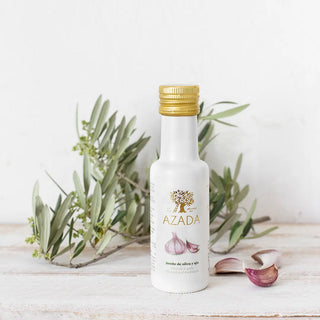 Fotografía frontal del aceite de ajo, acompañado de tres ajos frescos a su lado, con una rama de olivo de fondo que realza su carácter artesanal y mediterráneo. Real Fábrica.