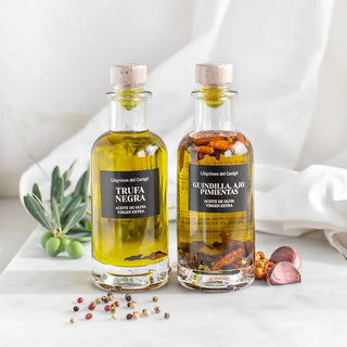 Foto de dos botellas de aceite de oliva virgen extra, una con trufa negra y otra con guindilla, ajo y pimientas, acompañadas de ingredientes frescos que resaltan su carácter artesanal y sabor intenso. Real Fábrica.