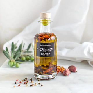 Foto de aceite de oliva virgen extra con guindilla, a su alrededor dos ajo y pimientas y una rama de olivo en el fondo. Real Fábrica