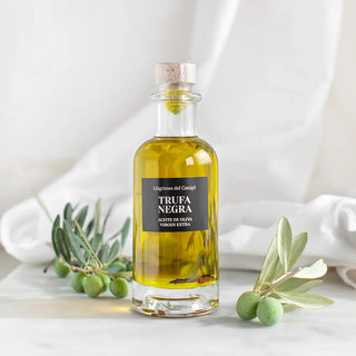 Foto de una botella elegante de aceite de oliva virgen extra de trufa negra, acompañada de ramas y aceitunas frescas, destacando su sabor premium y natural. Real Fábrica.