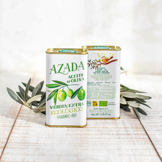 Foto frontal del aceite de oliva ecológico, mostrando el texto del frente y de la parte trasera del envase, con una rama de olivo al fondo que realza su carácter artesanal y mediterráneo. Real Fábrica.