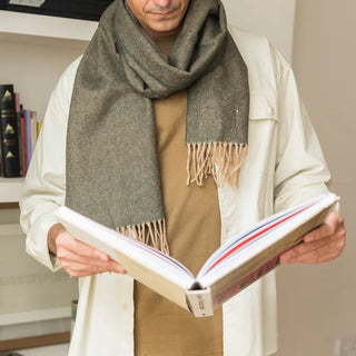 Foto de hombre luciendo en su cuello nuestra bufanda verde, mientras lee un libro. nuestras bufandas de lana merino, con un tejido de alta calidad, suave, fino y transpirable.  Real Fábrica.