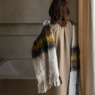 Una mujer de espalda luciendo nuestra bufanda de mohair tartán verde, negro y amarillo que lleva al cuello, una fibra suave y de gran calidad desarrollada por Ezcaray, en la Rioja. Real Fábrica.