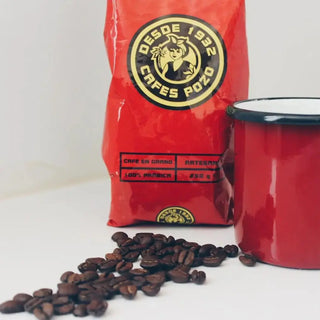 Foto que muestra el Café Pozo con su logo amarillo y negro, acompañada por una taza de peltre roja con borde negro, y unos granos de café esparcidos frente a ellos sobre una superficie blanca, transmitiendo un estilo simple y artesanal. Real Fábrica.
