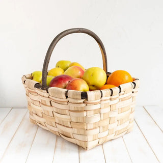 Foto de Esparraguera de castaño con asa con manzanas rojas y amarillas en su interior sobre una mesa de madera. Real Fábrica.