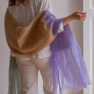 Mujer de espalda luciendo nuestro Chal bufanda de mohair en 3 colores: lavanda, camel y gris perla. En la imagen se aprecian en detalle sus terminaciones en largos flecos finos, hilados a mano. Real Fábrica.
