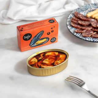 Foto muestra la conserva abierta con el producto al lado un tenedor y un plato con salame cortado y picos sobre una mesa blanca. Real Fábrica.