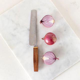 Vista cenital de cuchillo cebollero con 3 cebollas rojas sobre una tabla de corte de mármol.  mango de madera de acacia con filo dorado y acero de primera calidad. Elaborados artesanalmente en Galicia. Real Fábrica.