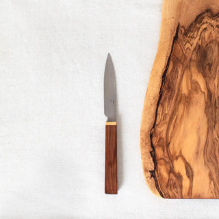 Vista cenital de cuchillo patatero junto a una tabla de corte de madera de boj.  mango de madera de acacia con filo dorado y acero de primera calidad. Elaborados artesanalmente en Galicia. Real Fábrica.