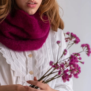 Mujer abrigada con un suave y esponjoso cuello de mohair. Sujeta un ramillete de flores del mismo color que el mohair, buganvilla. Real Fábrica.