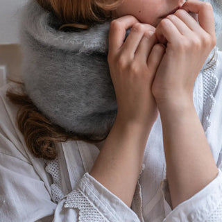 Mujer agarrando un cuello de mohair con ambas manos. La fibra es de color gris perla y en la imagen se observa la calidad del tejido artesano. Real Fábrica.