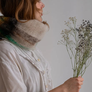 Mujer abrigada con un cuello de mohair con diseño de tartán, sujetando un ramillete de flores. El cuello está confeccionado a mano en La Rioja. Real Fabrica.