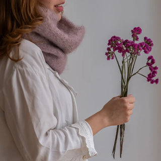 Mujer con un ramillete de flores en la mano y abrigada con un cuello de mohair en color violeta rosado. Real Fábrica.