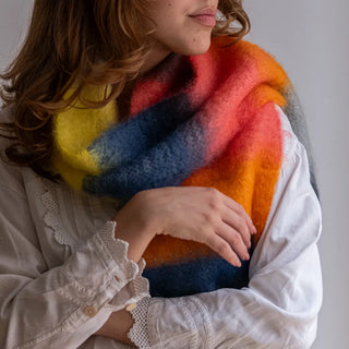 Foto de mujer luciendo nuestro foulard de mohair de franjas naranjas, rosas y berenjena, alrededor del cuello. El mohair es una fibra de gran calidad, suave y abrigada. Real Fábrica.