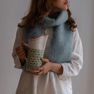 Mujer sujetando una jarra de cerámica de Cáceres con bordes carmín y con un foulard de mohair gris celeste alrededor del cuello. Real Fábrica.