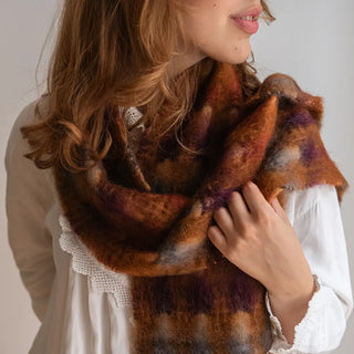 Mujer posando con un foulard de mohair con cuadros marrones y gris azulón. Acaricia con la mano la fibra suave y ligera. Real Fábrica.