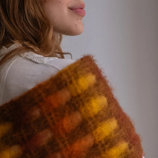 Foto de mujer luciendo un foulard de mohair sobre el hombro. Tiene un diseño de cuadros naranja y mostaza. Real Fábrica.