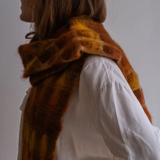 Mujer abrigada con un foulard de cuadros naranja y mostaza. Es de mohair, una fibra cálida y suave, elaborado a mano en La Rioja. Real Fábrica.