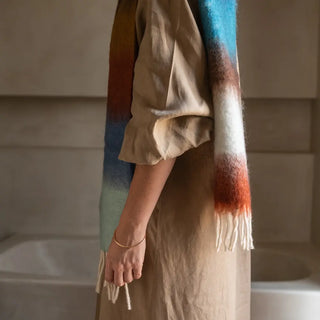 Foto mujer de perfil luciendo un foulard de mohair de franjas azul, mostaza y blanco que lleva en su hombro, una fibra suave y de gran calidad desarrollada por Ezcaray, en la Rioja. Real Fábrica.