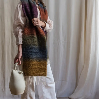 Mujer con una bufanda ancha hecha de mohair con un patrón de franjas de distintos colores oscuras. Sujeta un botijo.