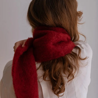 Mujer de espaldas con un foulard de mohair envolviendo su cuello. Toca con su mano la suave fibra de mohair en color rojo oscuro. Real Fábrica.