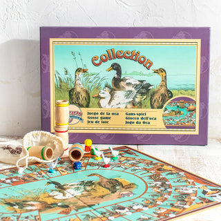 Plano frontal del juego de la oca. Al fondo, caja de cartón en vertical, mostrando la portada con ilustraciones antiguas. En primer plano, sobre una mesa de madera blanca, un escorzo del tablero abierto en el que se encuentran las fichas del juego. Real Fábrica.