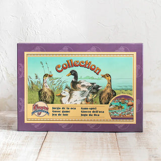 Caja de cartón duro del tradicional juego de la oca, con ilustraciones en estilo antiguo, sobre una mesa de madera blanca. Real Fábrica.