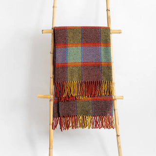 Manta riojana de lana de cuadros en tonos verde y rojo con flecos colgada de una escalera de madera vintage. Real Fábrica