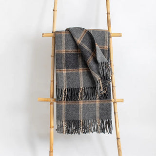 Manta riojana de lana de cuadros en tonos gris y beige con flecos colgada de una escalera de madera vintage. Real Fábrica