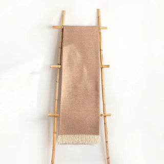 Manta en color camel expuesta en una escalera de madera con una textura de espiga. Flecos anudados a mano, uno a uno. Real Fábrica.
