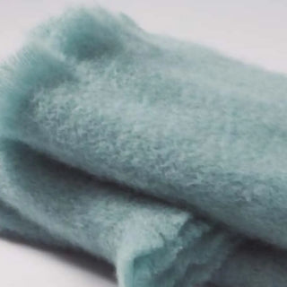 Detalle de manta de mohair en color gris celeste, con terminación en flecos finos. En el primer plano se aprecia la textura de la fibra de mohair. Real Fábrica.