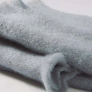 Detalle de manta de mohair en color gris perla, con terminación en flecos finos. En el primer plano se aprecia la textura de la fibra de mohair. Real Fábrica.