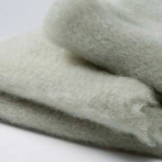 Detalle de manta de mohair en color gris verde, con terminación en flecos finos. En el primer plano se aprecia la textura de la fibra de mohair. Real Fábrica.