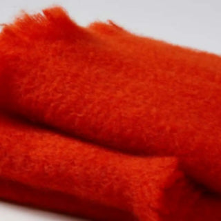 Detalle de manta de mohair en color naranja oscuro, con terminación en flecos finos. En el primer plano se aprecia la textura de la fibra de mohair. Real Fábrica.