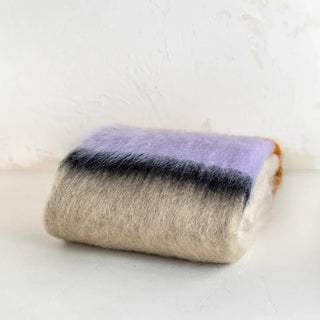 Manta de mohair cuidadosamente doblada. Tiene un diseño de rayas lilas, negras y marrones. Real Fábrica.