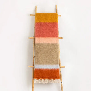 Manta expuesta sobre una escalera vintage, esta manta de rayas en tonos terracota y caldera, elaborada artesanalmente en mohair de altísima calidad. Real Fábrica.