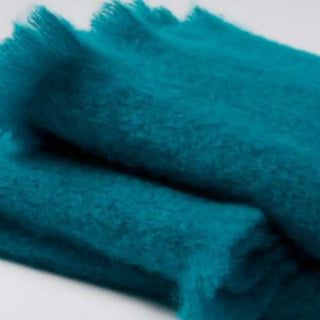 Detalle de manta de mohair en color turquesa, con terminación en flecos finos. En el primer plano se aprecia la textura de la fibra de mohair. Real Fábrica.