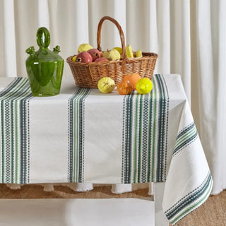 Mantel de rayas verdes, blancas y negras con un botijo de cristal verde y un cesto lleno de frutas, ideal para una mesa fresca y natural. Real Fábrica.