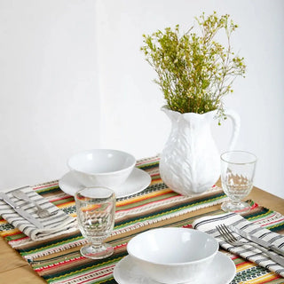 Foto de dos manteles individual alpujarra tonos cálidos y vajilla de cerámica blanca cubiertos, vasos y un jarrón con flores, en un ambiente acogedor. Real Fábrica.