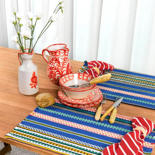 Mesa con dos manteles individual de rayas coloridas y vajilla de cerámica de granada roja, con jarra, cubiertos y pan rústico, en un ambiente acogedor. Real Fábrica.