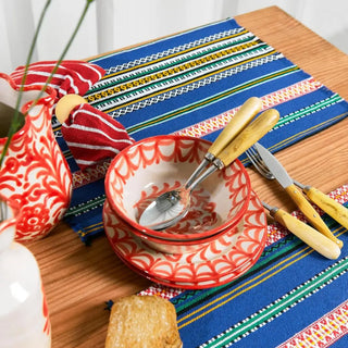 Foto cenital de un mantel individual de rayas coloridas y vajilla de cerámica de granada roja cubiertos y pan rústico, en un ambiente acogedor. Real Fábrica.
